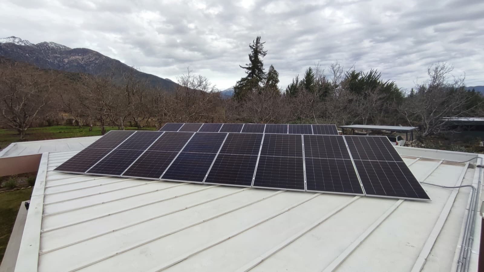 Paneles solares sobre un techo metálico bajo un cielo nublado de invierno, con la cordillera nevada al fondo