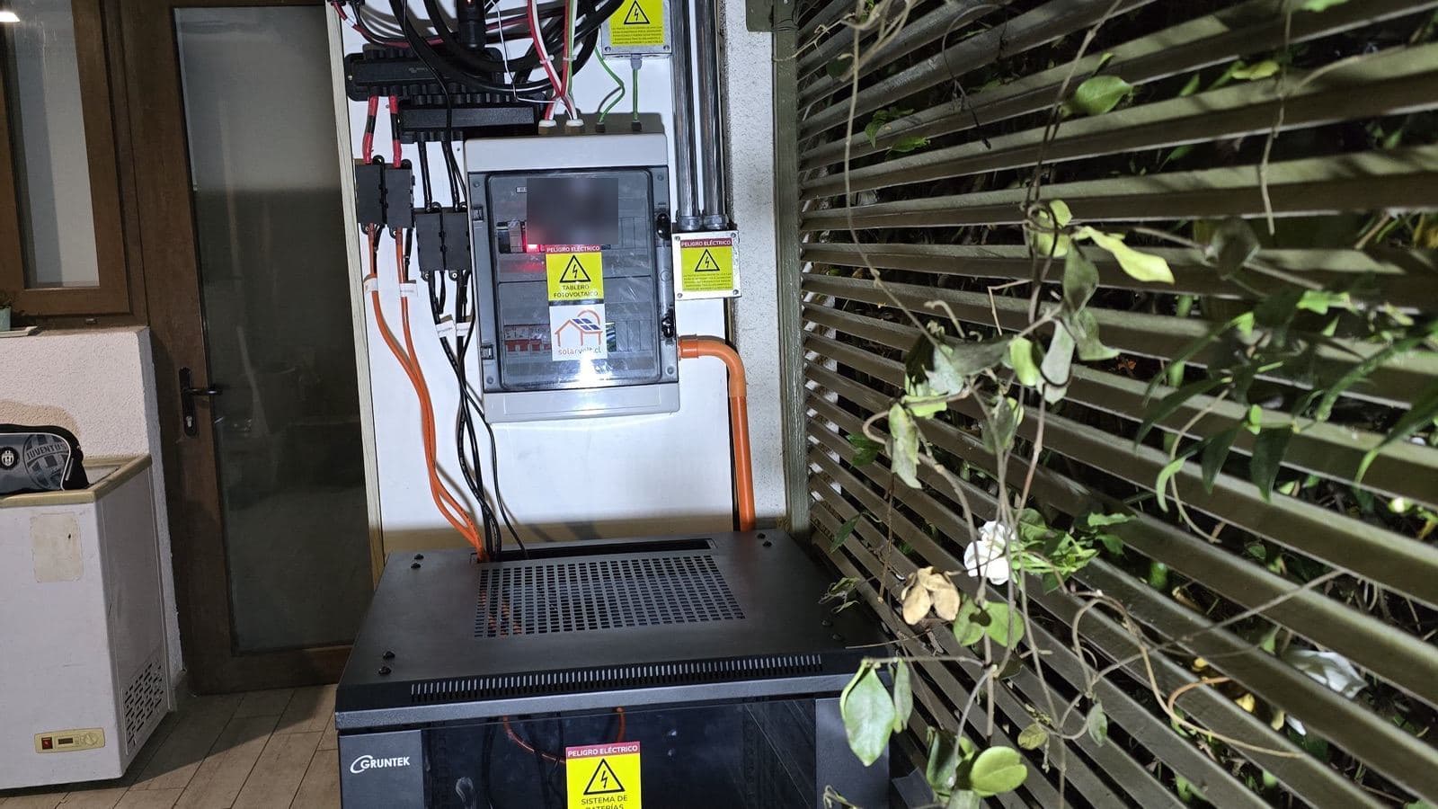 Tablero fotovoltaico con letreros de peligro eléctrico, cables y un gabinete de baterías, junto a una celosía de madera con enredadera