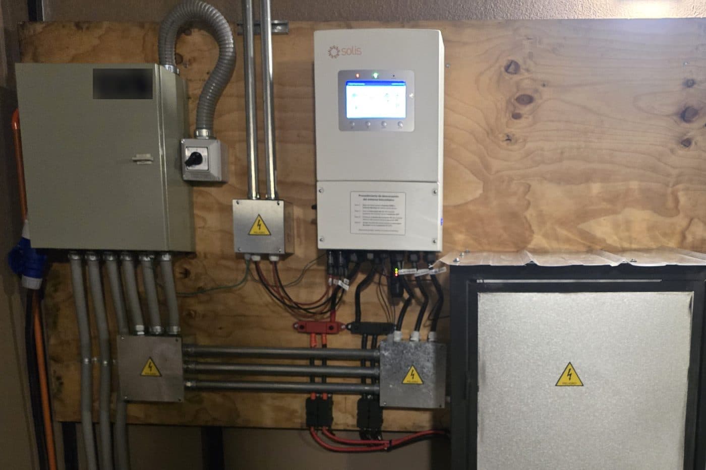 Inversor blanco con pantalla montado en un muro de madera, junto a un tablero eléctrico gris, cajas de conexión con señal de peligro y canalizaciones metálicas