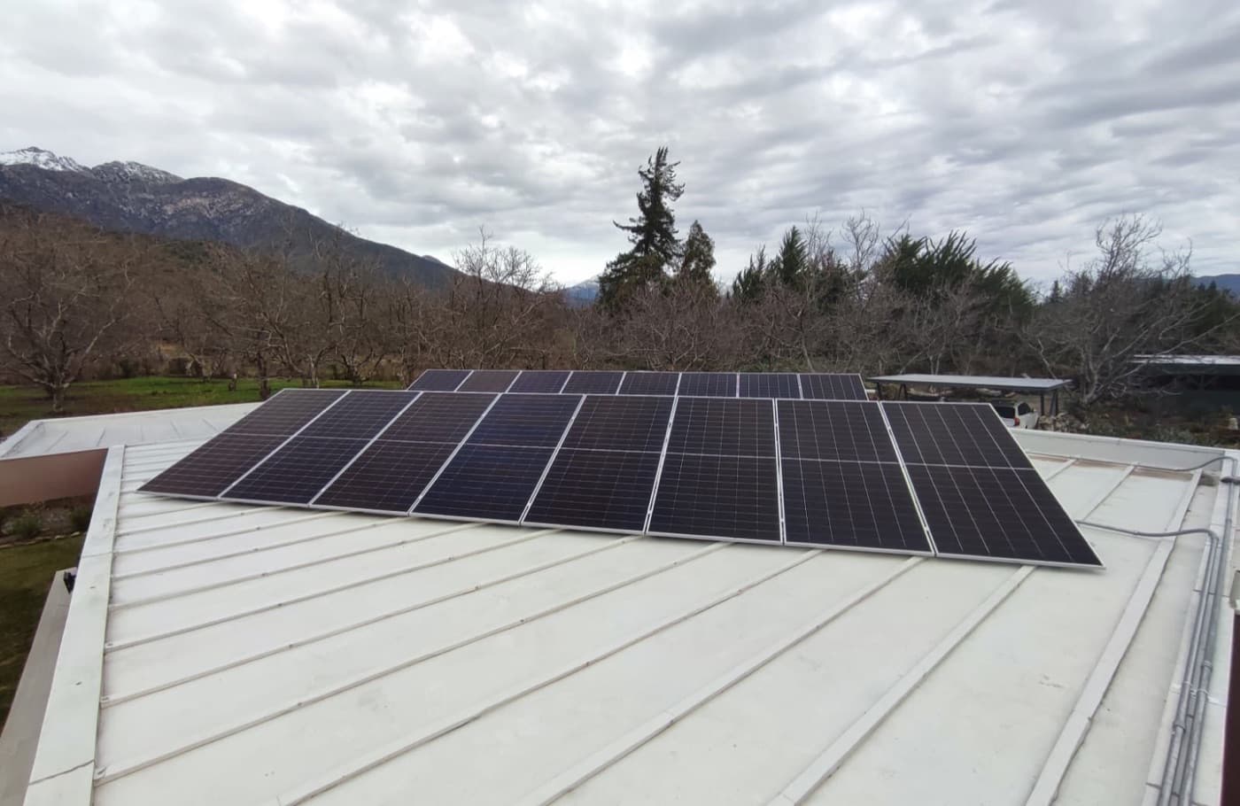Paneles solares sobre un techo metálico blanco, bajo un cielo nublado de invierno con la cordillera al fondo