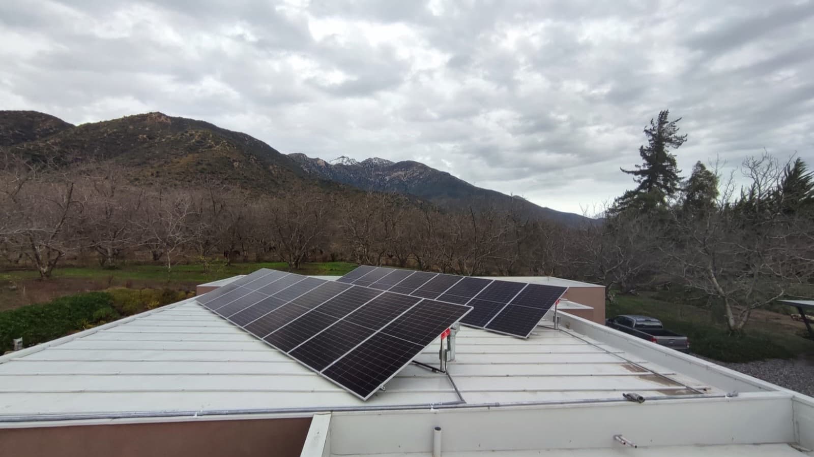 Paneles solares inclinados sobre el techo metálico plano de una casa rural, con un huerto sin hojas y la cordillera nevada bajo un cielo nublado