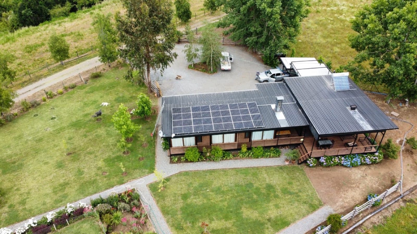Vista aérea de una casa de campo de madera con techo negro y paneles solares, rodeada de césped y árboles