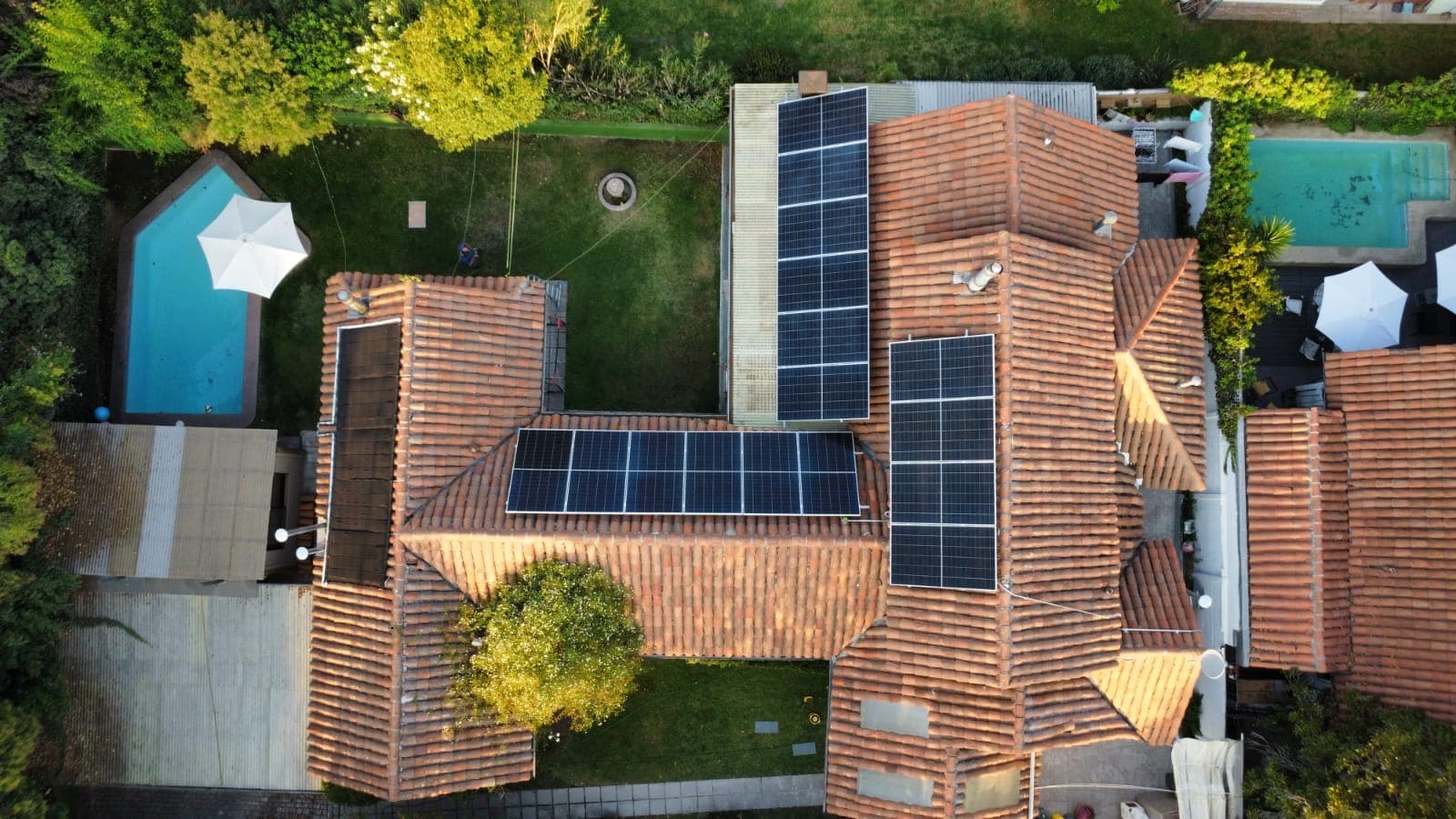 Vista aérea de una casa con techo de tejas de arcilla y paneles solares en varias de sus aguas, junto a un jardín y dos piscinas