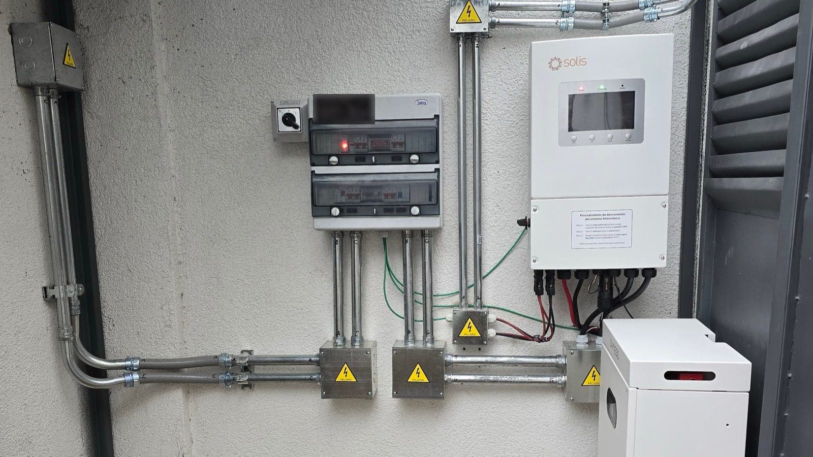 Inversor, tablero fotovoltaico y cajas de conexión montados en un muro exterior, con un módulo de baterías al costado